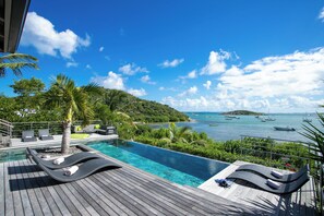 Un espace aménagé au bord de la piscine, avec des chaises longues, un palmier et une vue sur la mer où l’on peut apercevoir des bateaux.