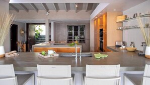A modern kitchen with a central island, built-in appliances, and a dining area with white chairs. - Luxury Waterfront Villa Pinel. Direct Ocean Views & Private Beach Access (SAINT MARTIN)