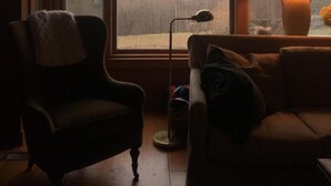 Una habitación con un ventanal que ofrece vistas a un paisaje boscoso. Hay una silla y una mesa con una lámpara.