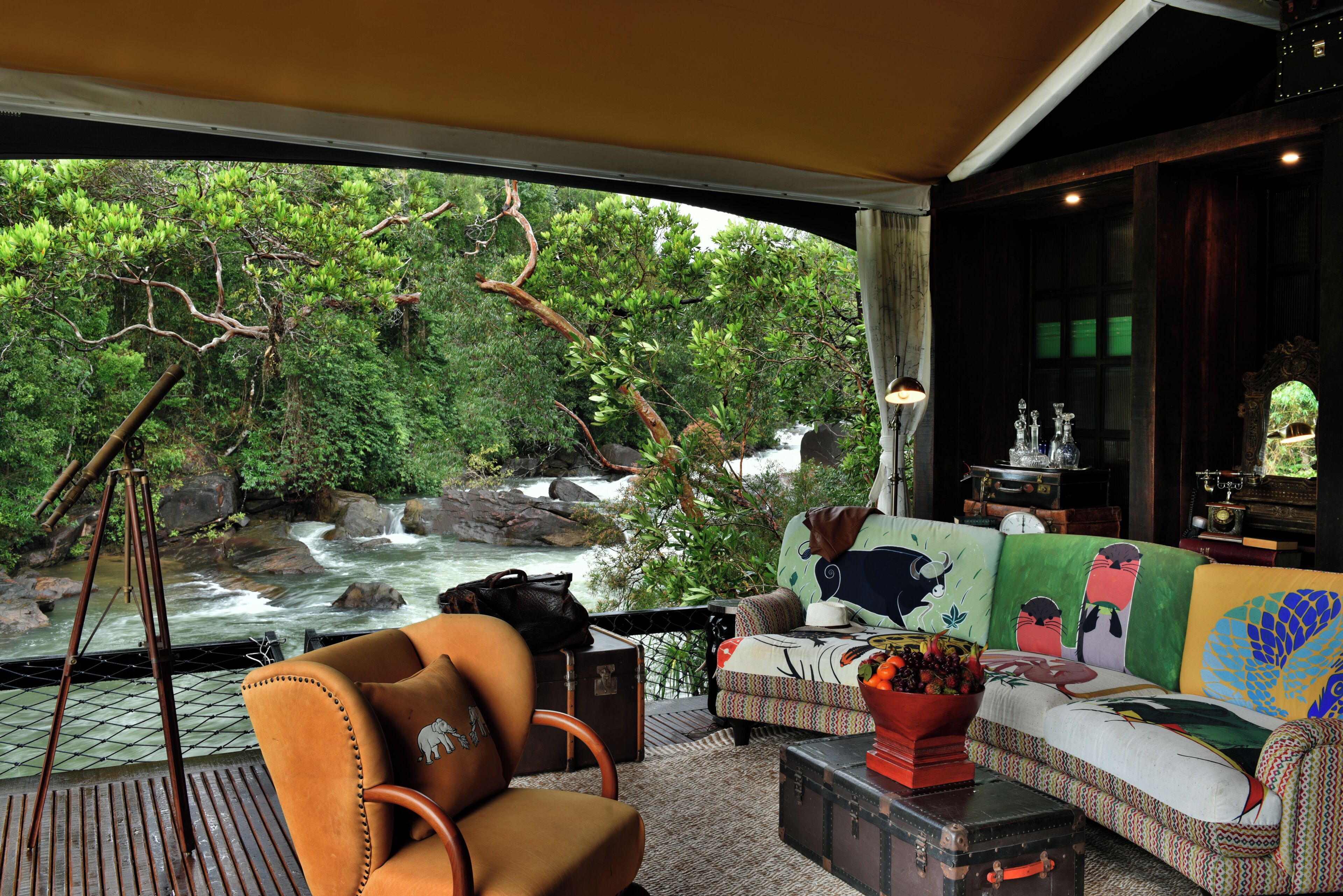Habitación con sofá mirando a una ventana con vista al río, un trípode y una mesa con un jarrón de flores.