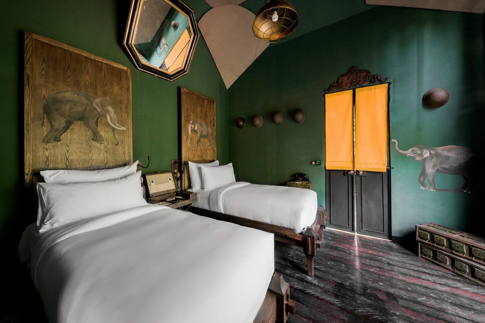 A room with two beds, a mirror, a lamp, and a wall with elephant artwork.
