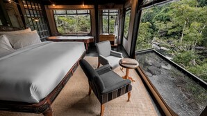 Une chambre à coucher avec un grand lit, une chaise, une petite table et une vue sur une forêt, visible à travers de grandes fenêtres.