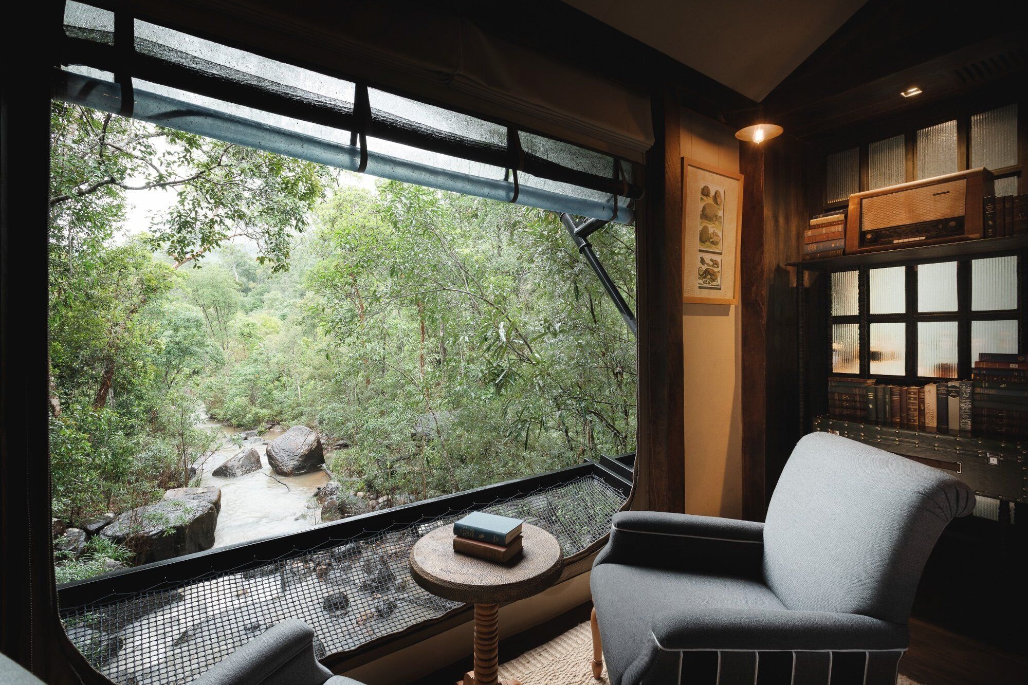 A room with a large window overlooking a forested area, a chair, a small table with a book, and a bookshelf with books.