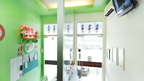 Un área de recepción con una pared verde, mostrador de recepción, un teléfono, una silla, felpudo de bienvenida y un televisor en la pared.
