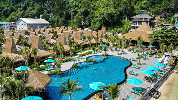 Un complexe hôtelier doté d’une grande piscine entourée de chaises longues et de parasols, d’une terrasse en bois et de structures à toits de chaume.