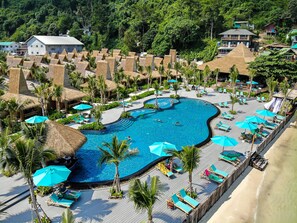 Ein Resort mit einem großen Schwimmbecken, umgeben von Liegestühlen und Sonnenschirmen, einer Holzterrasse und strohgedeckten Gebäuden.