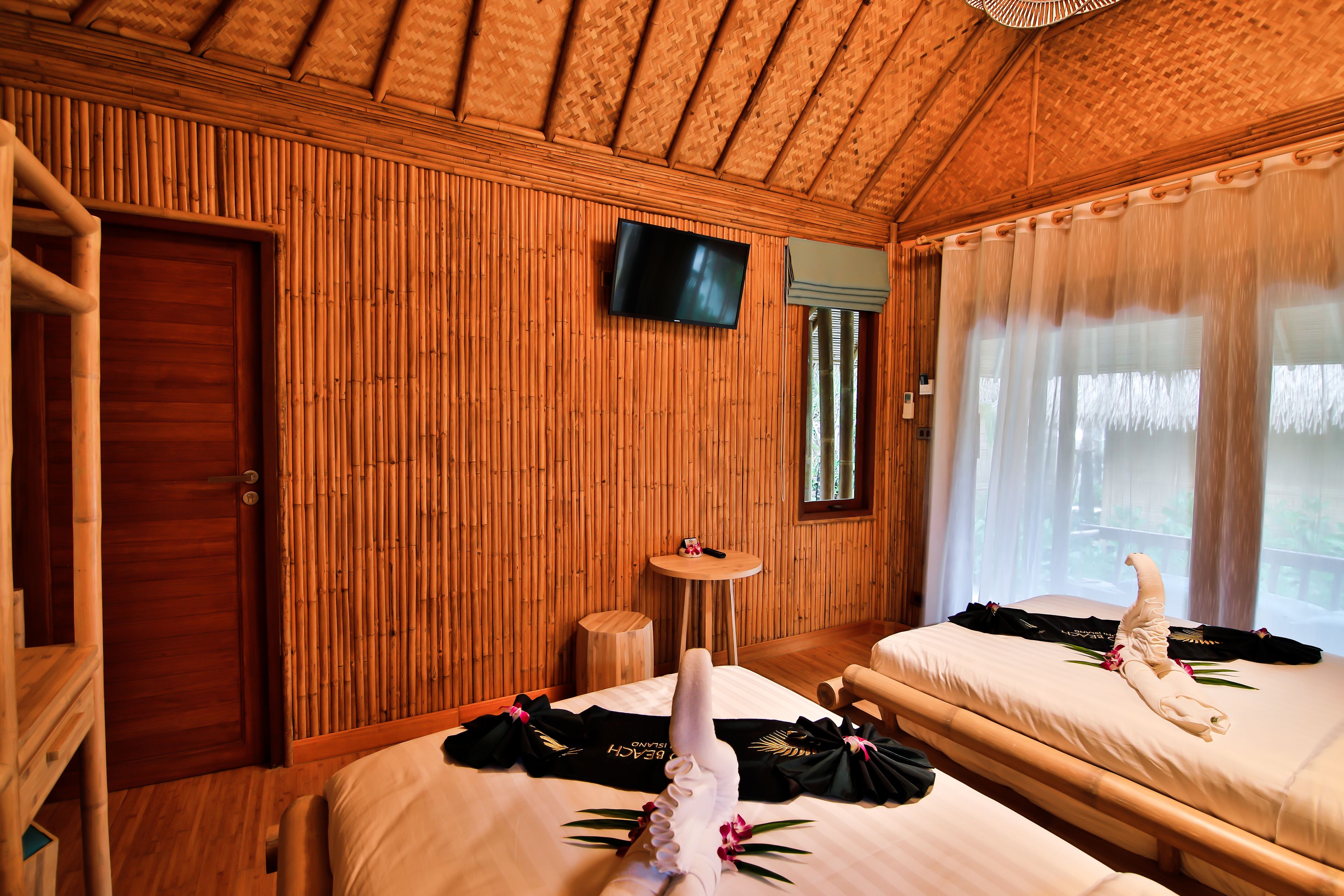 Una habitación con paredes de madera, un televisor montado en la pared, una cama con una toalla y un zapato, y una pequeña mesa redonda con un control remoto.