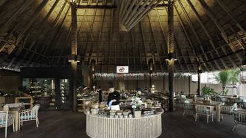 A restaurant with a high, thatched roof interior, wooden tables and chairs, and a central counter with staff.