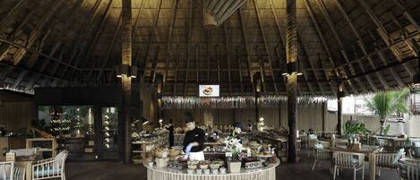 A restaurant with a high, thatched roof interior, wooden tables and chairs, and a central counter with staff.