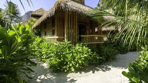 Un bungalow au toit de chaume, doté d’une véranda en bois, entouré d’une végétation tropicale luxuriante.
