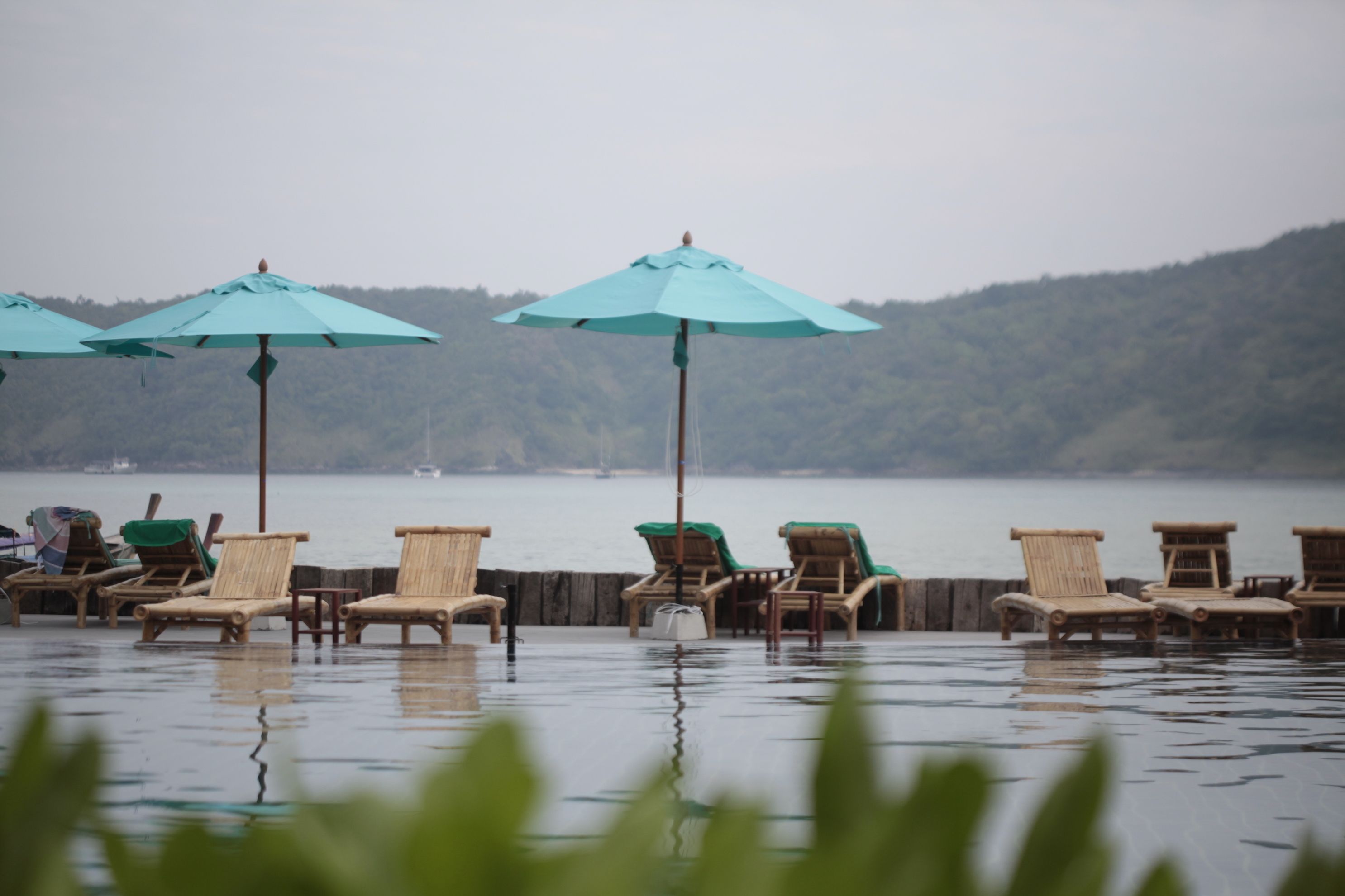 A waterfront area with wooden lounge chairs and turquoise umbrellas, overlooking a calm body of water and a forested hill.