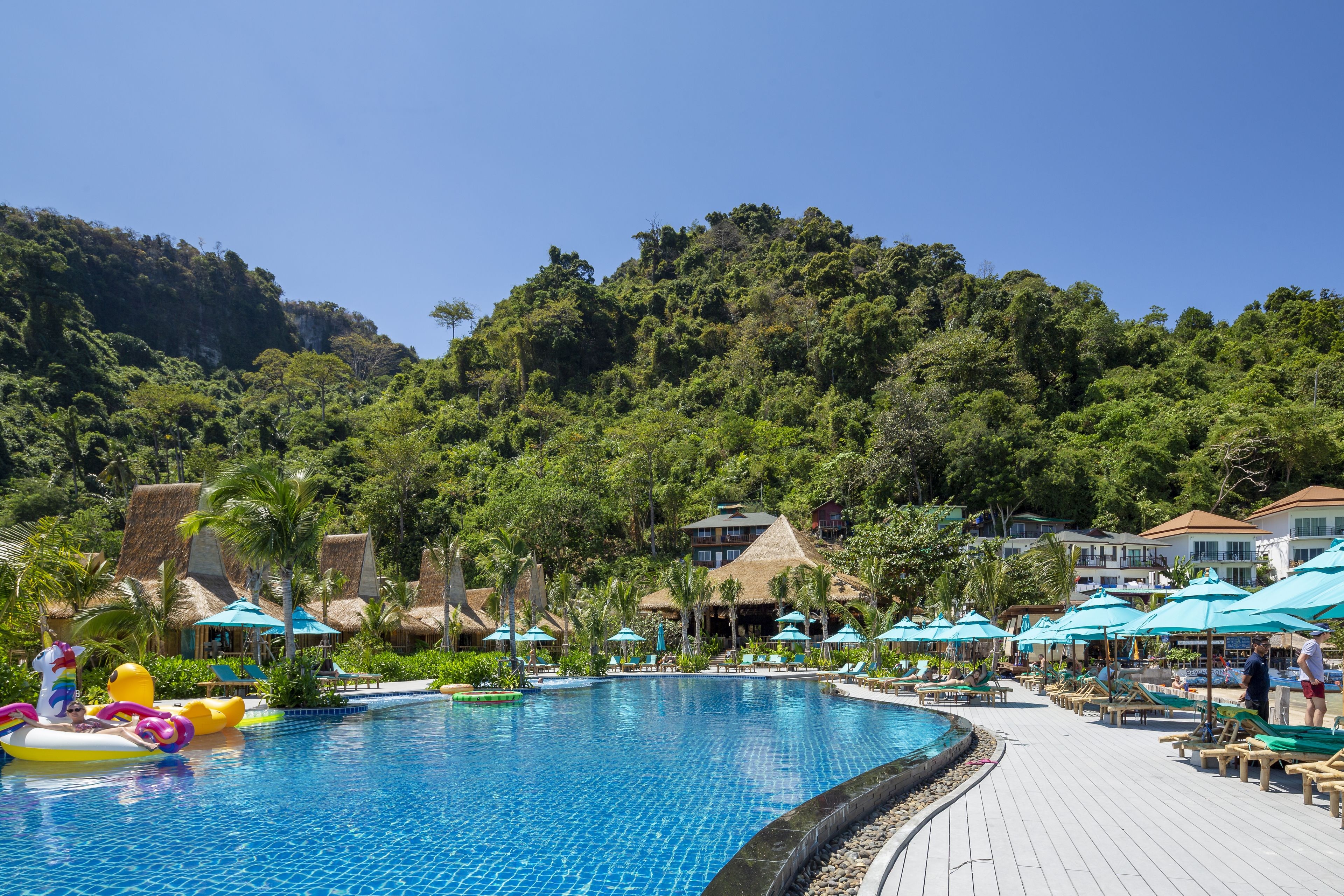 Ein Resort-Poolbereich mit einem klaren, blauen Pool, Liegestühlen, Sonnenschirmen und Gebäuden mit Strohdächern.