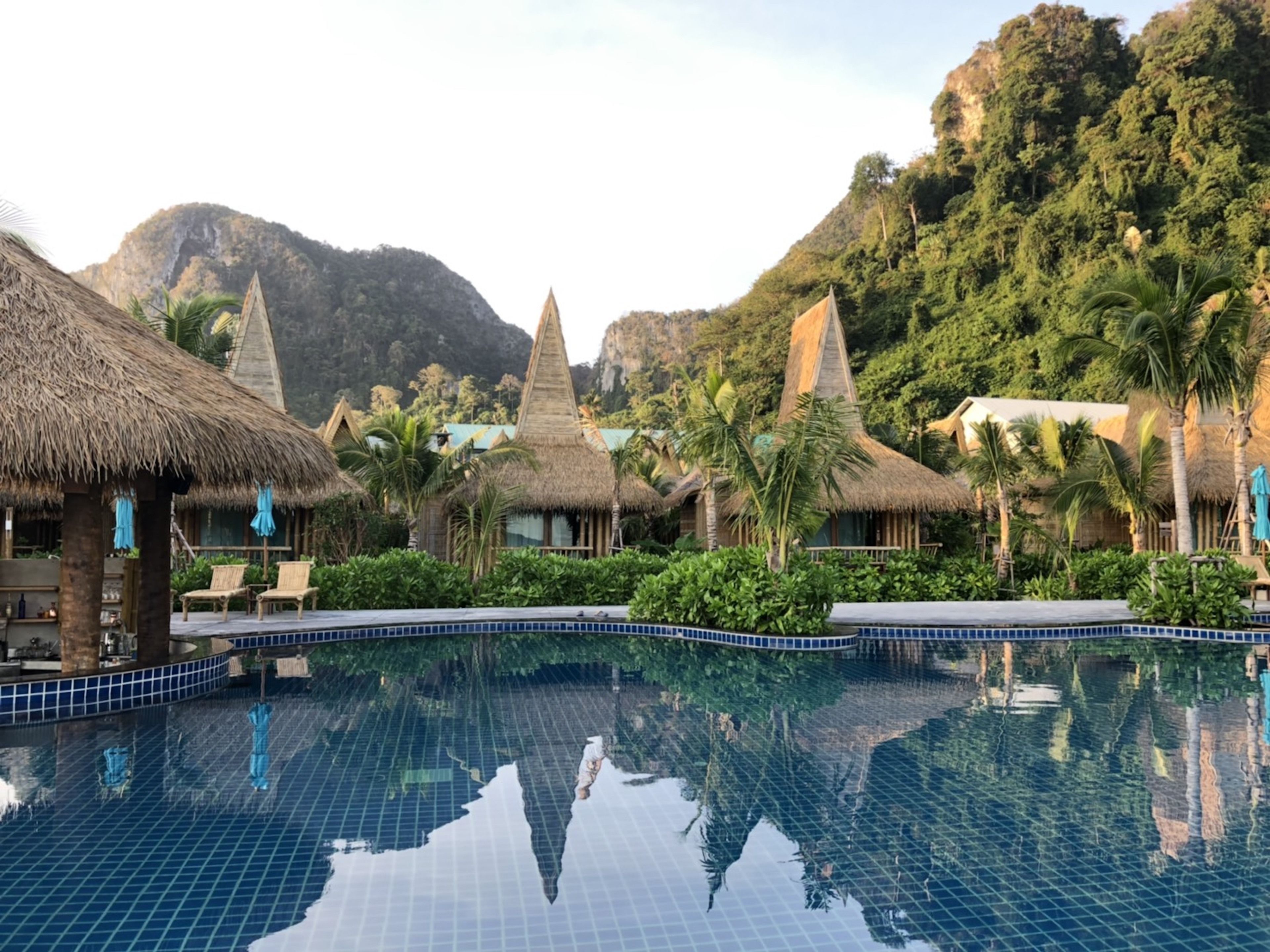 A pool with clear blue tiles, surrounded by thatched-roof structures and lush greenery, set against a mountainous backdrop.