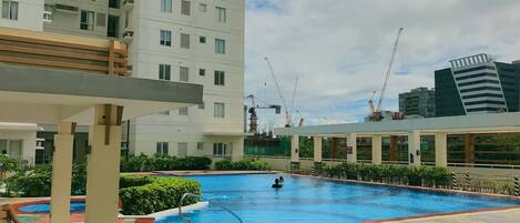 A swimming pool with a tiled deck, surrounded by greenery and modern buildings.