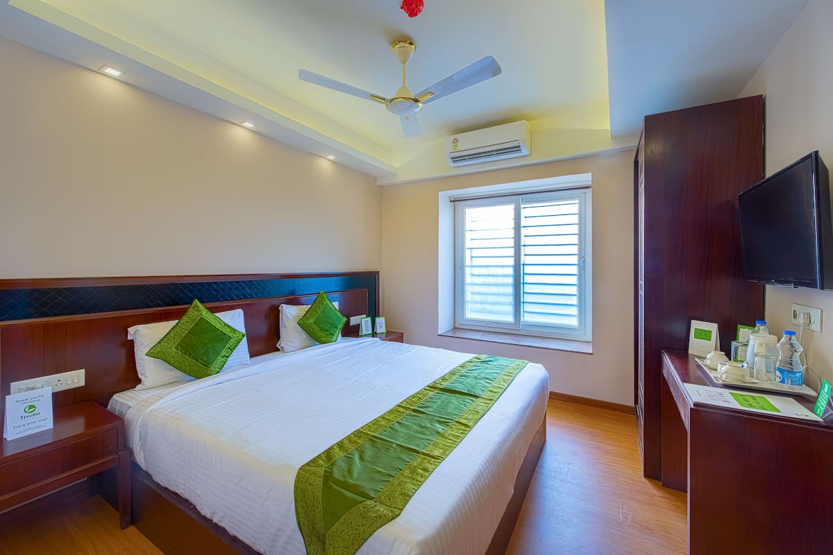 A hotel room with a large bed, a wooden headboard, a desk with a flat-screen TV, and a window with blinds.