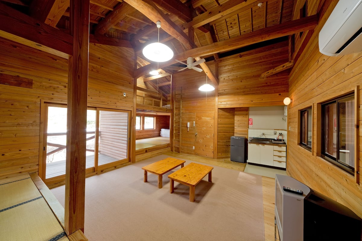 Interior de una cabaña de madera con suelo de esteras de tatami, dos mesas de madera, ventilador de techo y una pequeña cocina.