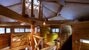 Interior de una cabaña con vigas de madera a la vista, un altillo y ventanas amplias.