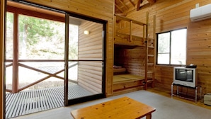 Interior de una cabaña de madera con un banco de madera, un televisor y un amplio ventanal corredera que da a un espacio exterior.