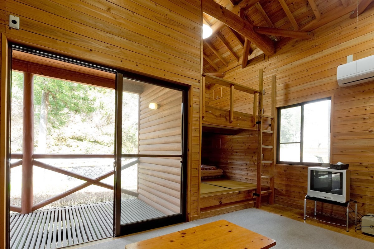 Interior de una cabaña de madera con un banco de madera, un televisor y un amplio ventanal corredera que da a un espacio exterior.