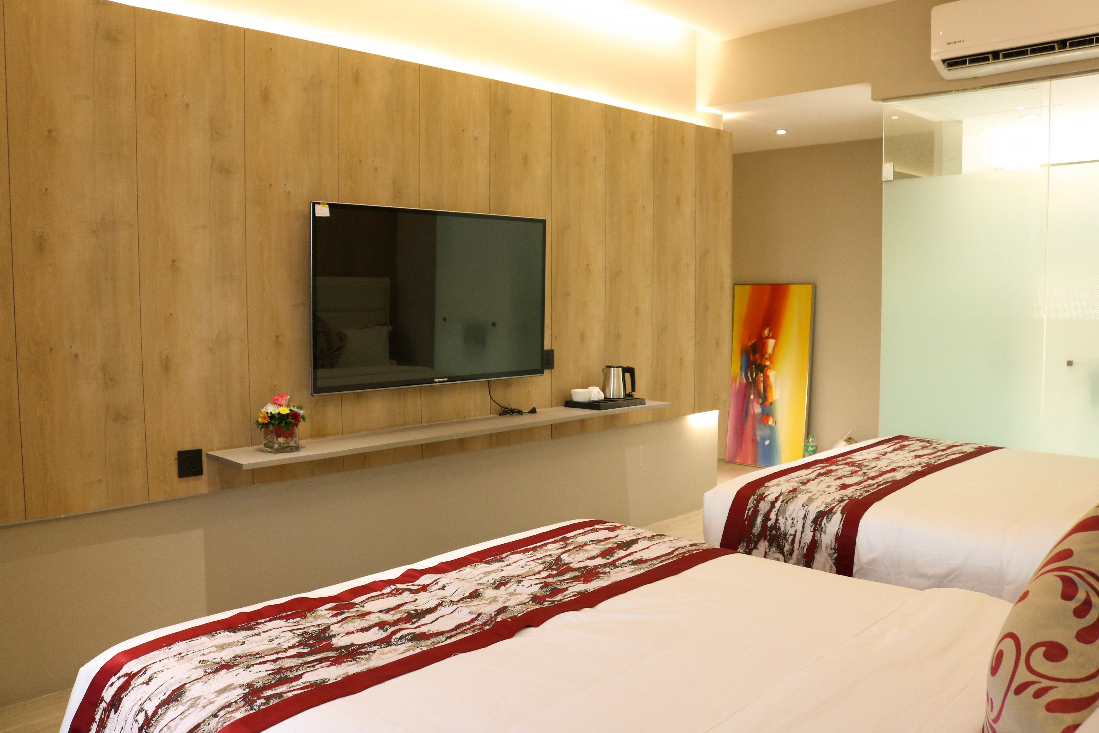 A hotel room with a wooden panel wall, a mounted flat-screen TV, a shelf with a coffee maker, and two beds with patterned bedspreads.