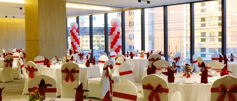 A banquet hall with round tables, white chairs with red sashes, and large windows offering a view of buildings.
