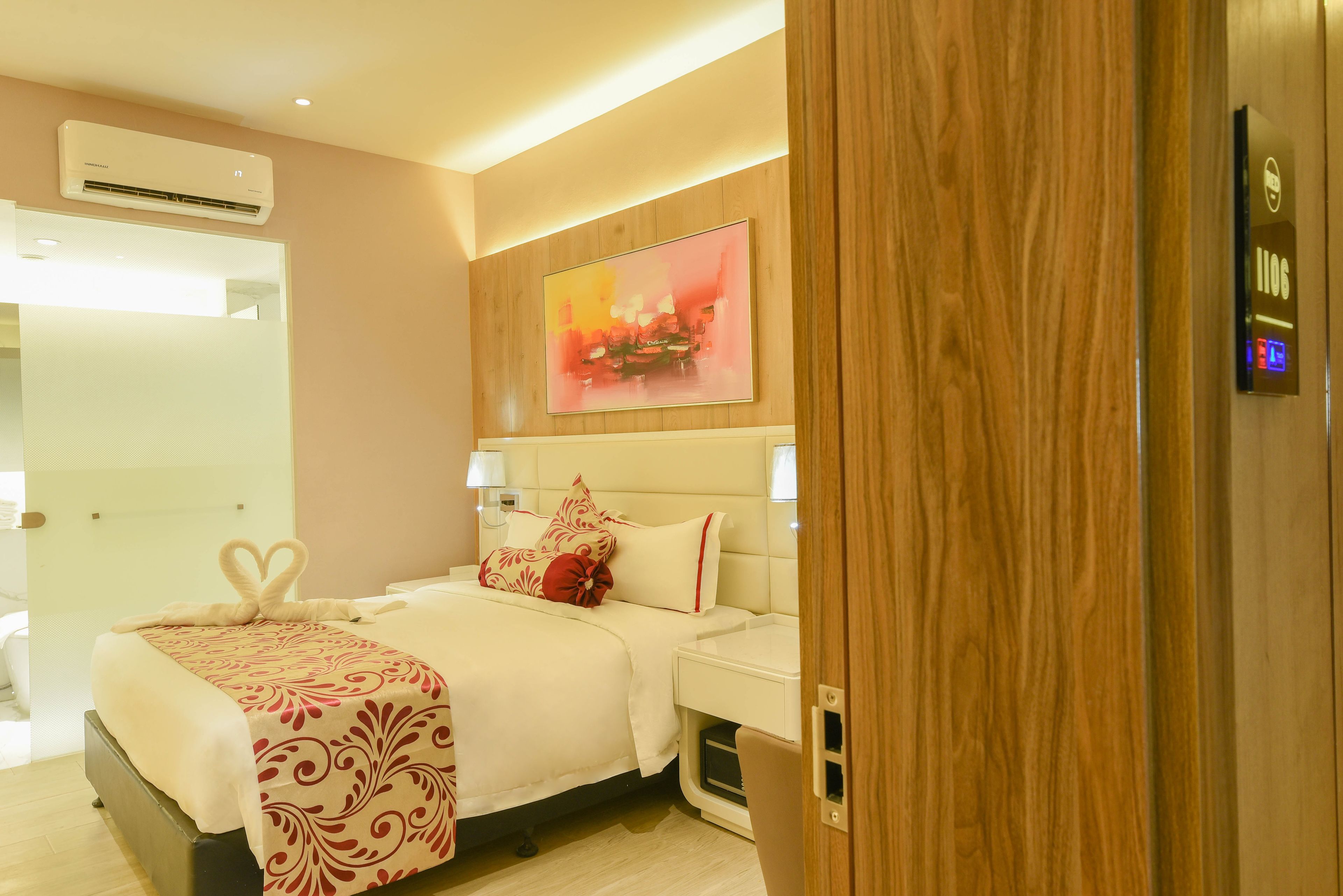 A modern hotel room with a large bed, a wooden panel wall, a painting, and a digital room service panel.
