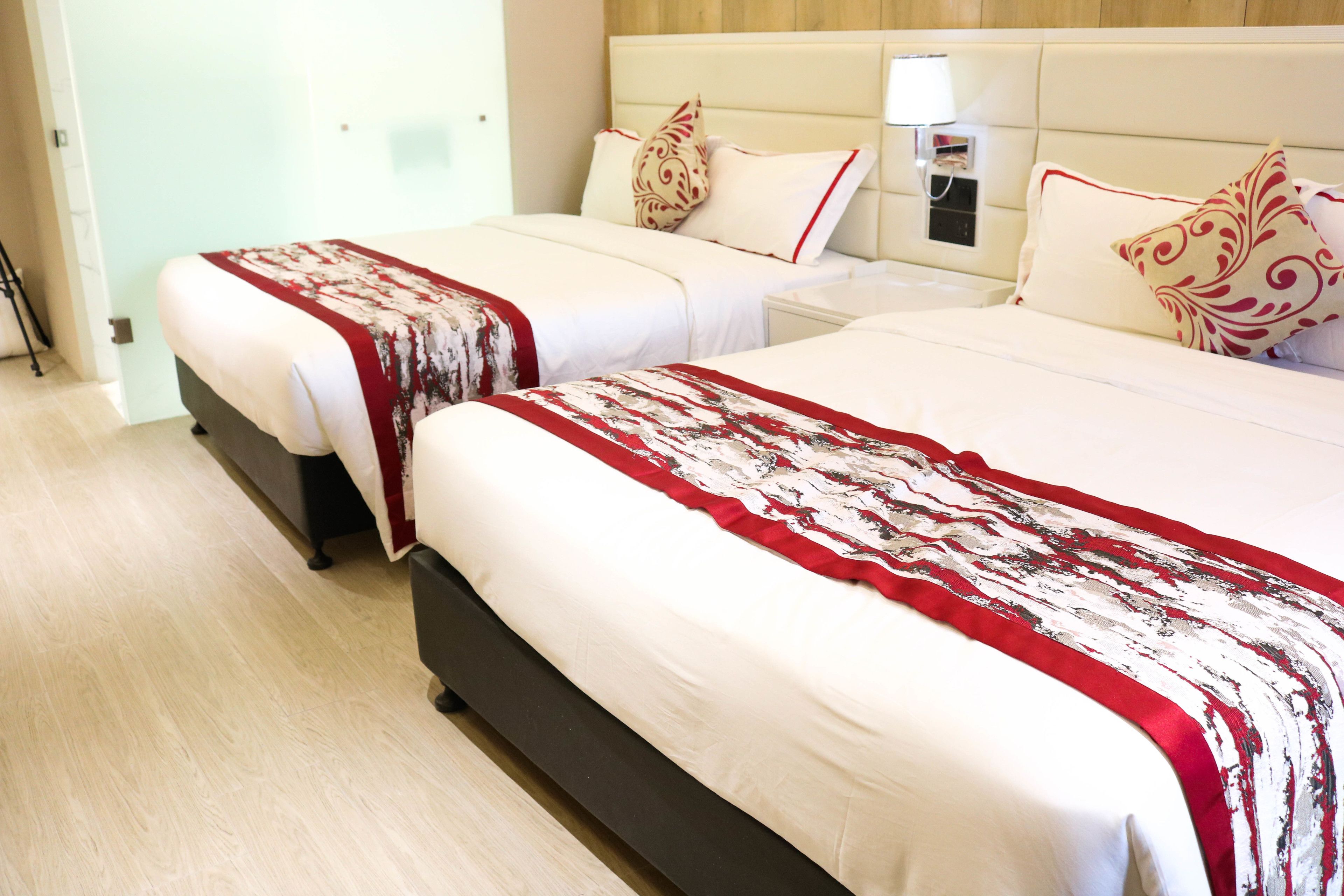 A hotel room with two beds, each with a red and white patterned bedspread, a wooden headboard, and a bedside table with a lamp.