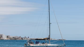 A sailboat with a tall mast and multiple sails, docked near a coastal cityscape with buildings in the background.