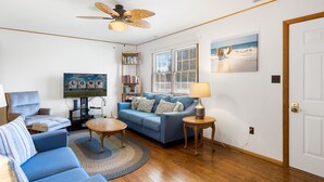 A living room with a blue sofa, a coffee table, a ceiling fan, a TV, a bookshelf, and a window with blinds. - Beach Pines by Seaside Vacations & Sales (Chincoteague Island)