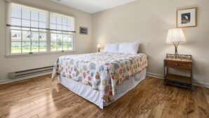 A bedroom with a large bed, a nightstand, a lamp, a framed picture on the wall, and a window with blinds. - Beach Pines by Seaside Vacations & Sales (Chincoteague Island)