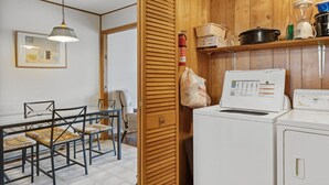 A compact kitchen with wooden cabinets, a washing machine, and a dining area with chairs and a table. - Beach Pines by Seaside Vacations & Sales (Chincoteague Island)