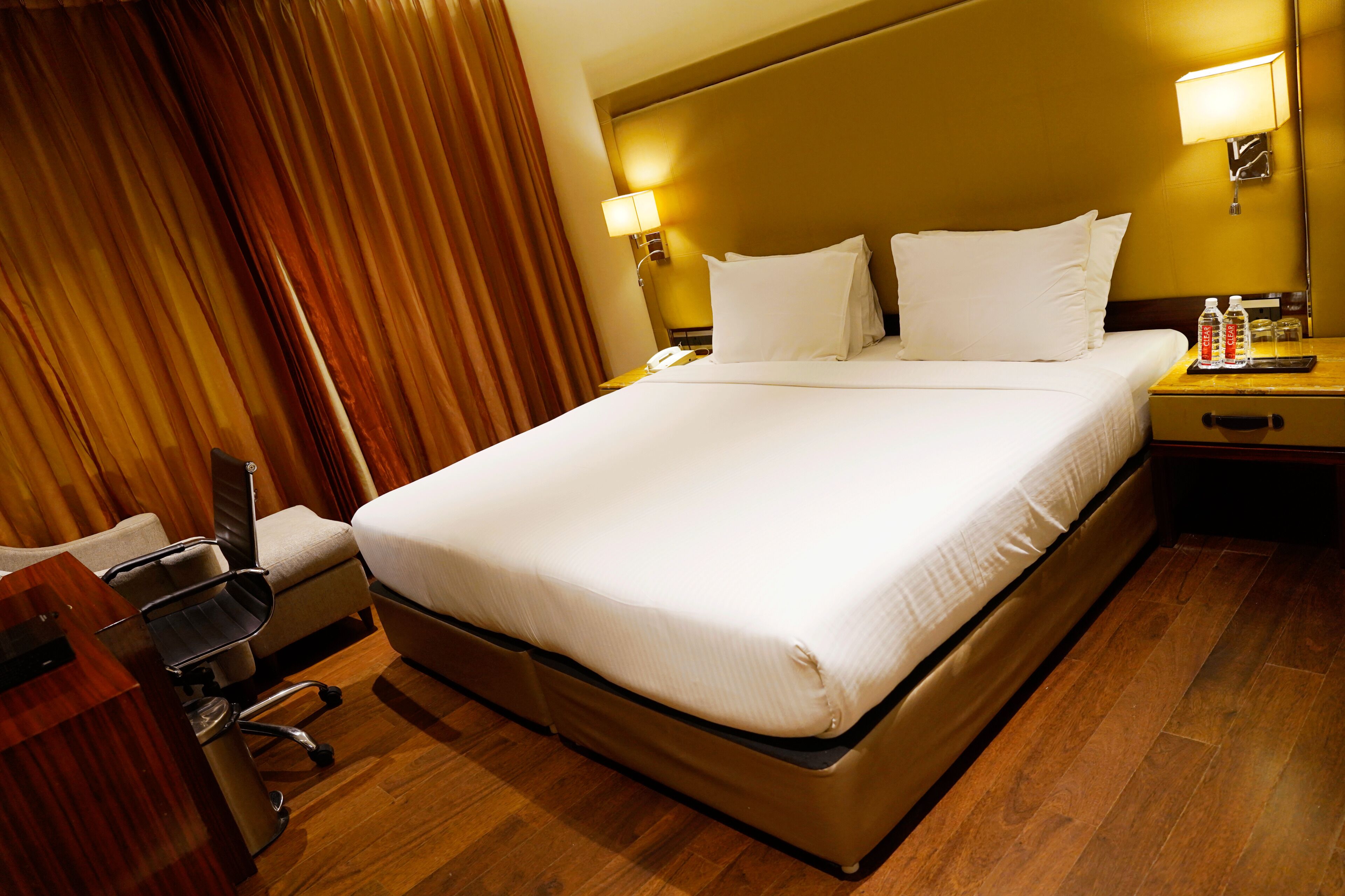 A hotel room with a large bed, a desk with a chair, a nightstand with a lamp, and a window with curtains.