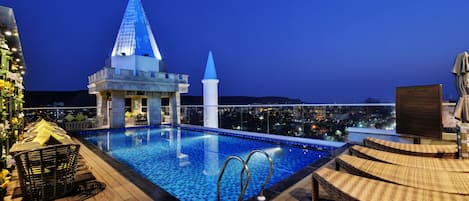 Una piscina en la azotea con vistas a la ciudad de noche, una estructura cónica y asientos tipo sala de estar.