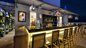 Un espace bar avec des fauteuils en osier, un comptoir avec des bouteilles et un mur orné d’un tableau encadré.