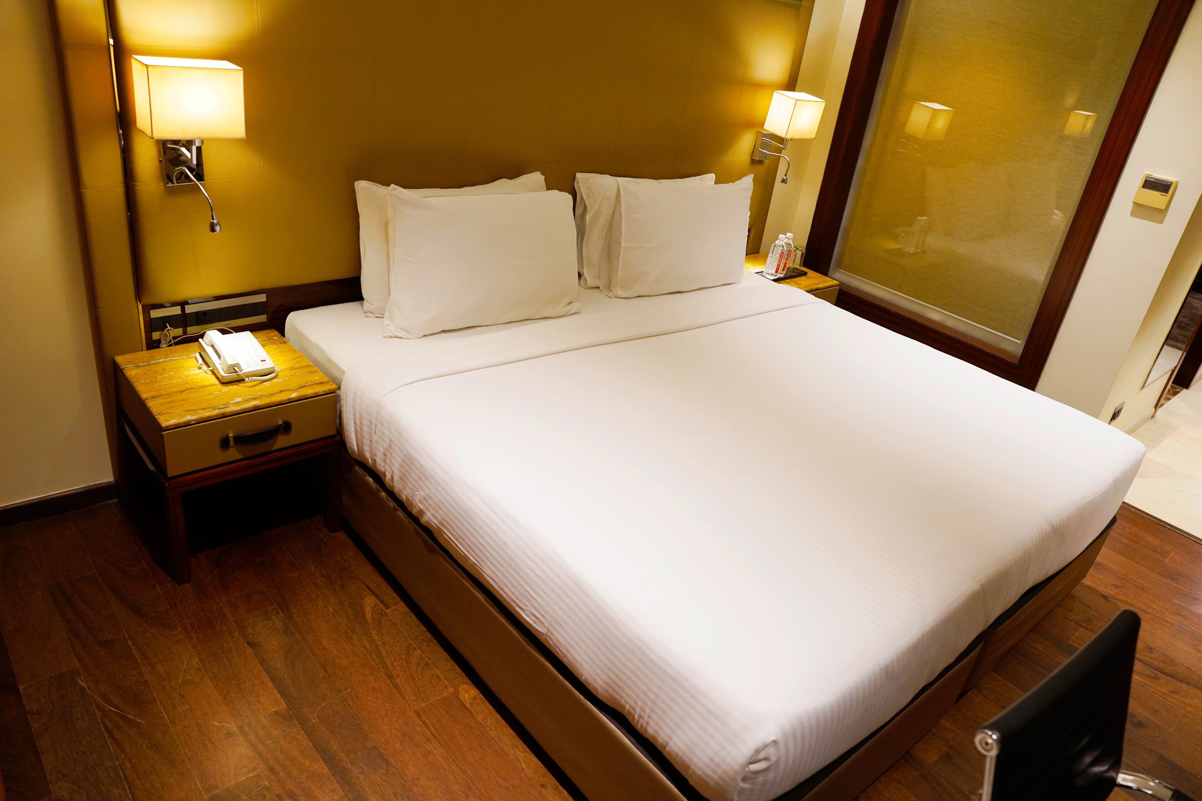 A hotel room with a large bed, two bedside lamps, a nightstand with a phone, and a wooden floor.