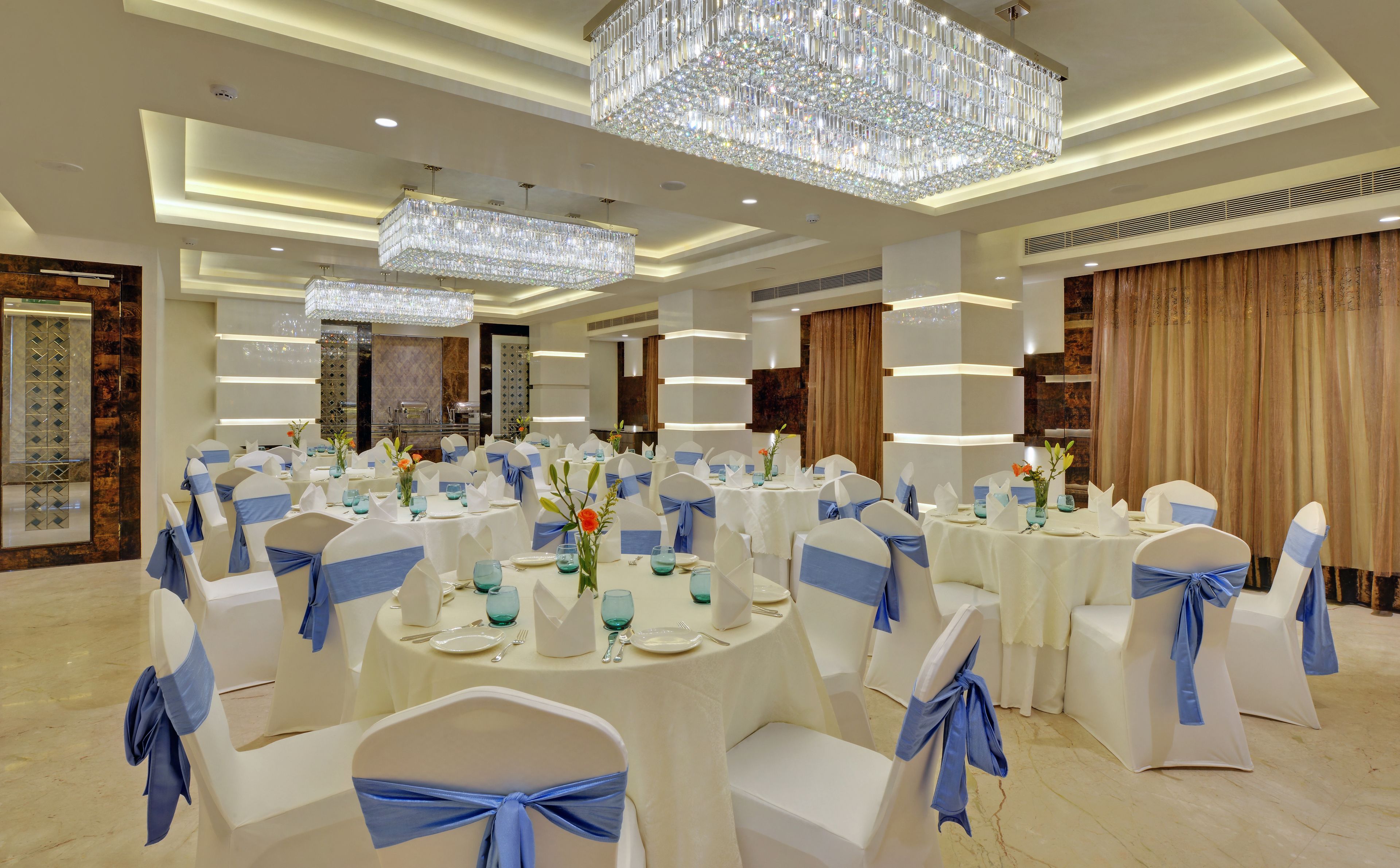 Une salle de banquet avec des tables rondes, des chaises blanches ornées de rubans bleus et un grand lustre en cristal.