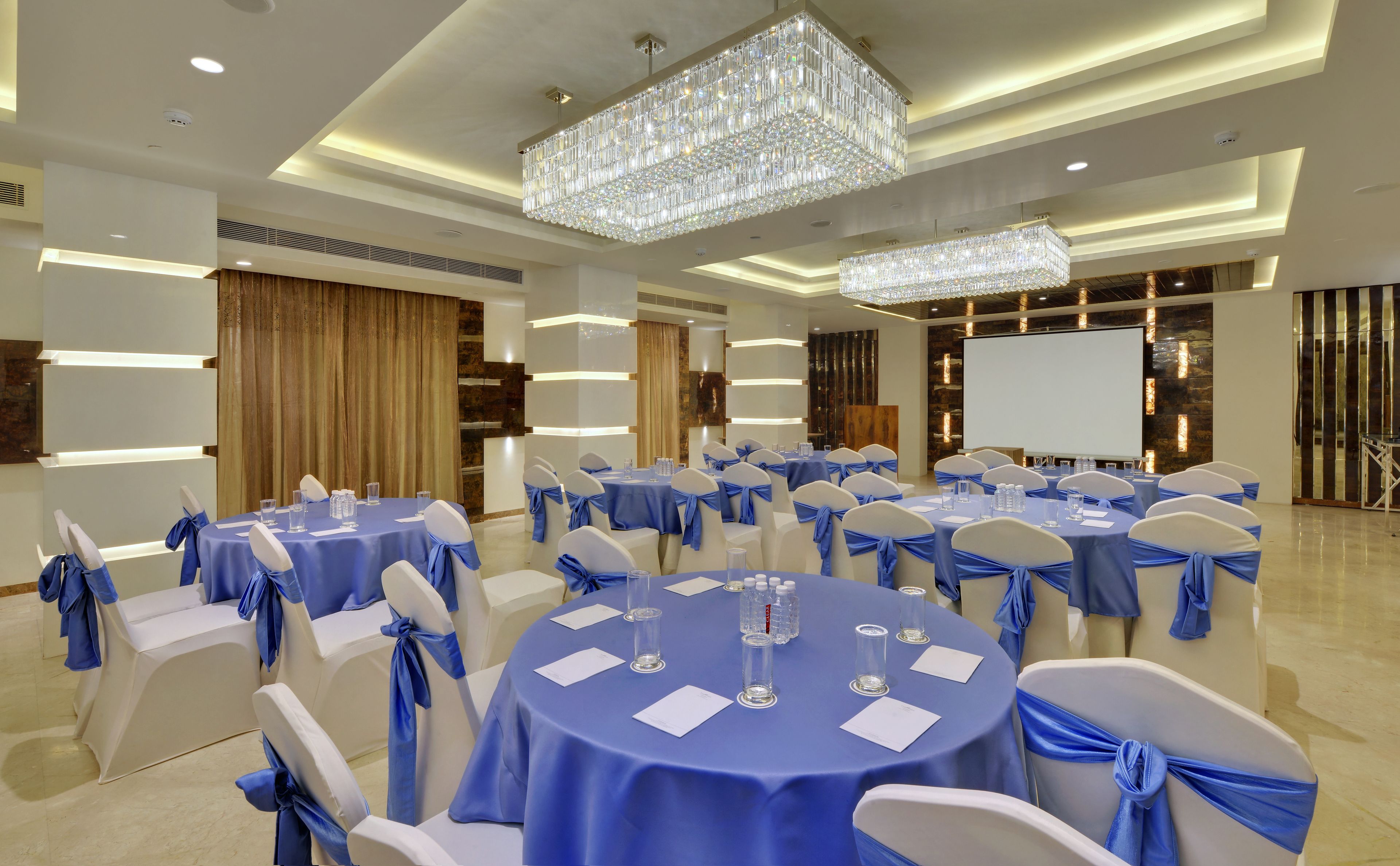 Un salón de conferencias con mesas redondas, manteles azules y sillas blancas adornadas con cintas azules.