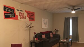 Een woonkamer met een zwarte leren bank, een rode fauteuil, een bijzettafeltje en een vloerlamp. Op de muur hangen posters en er is een plafondventilator.