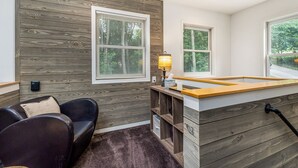 A cozy room with a wooden wall, a black leather sofa, a small wooden table with a lamp, and a window with blinds.