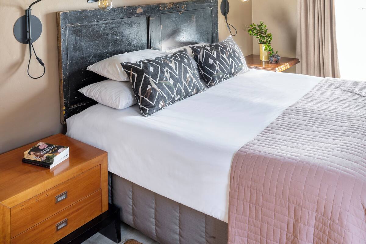 A bedroom with a bed, two bedside tables, a world map painting, a lamp, a plant, and a window with curtains.