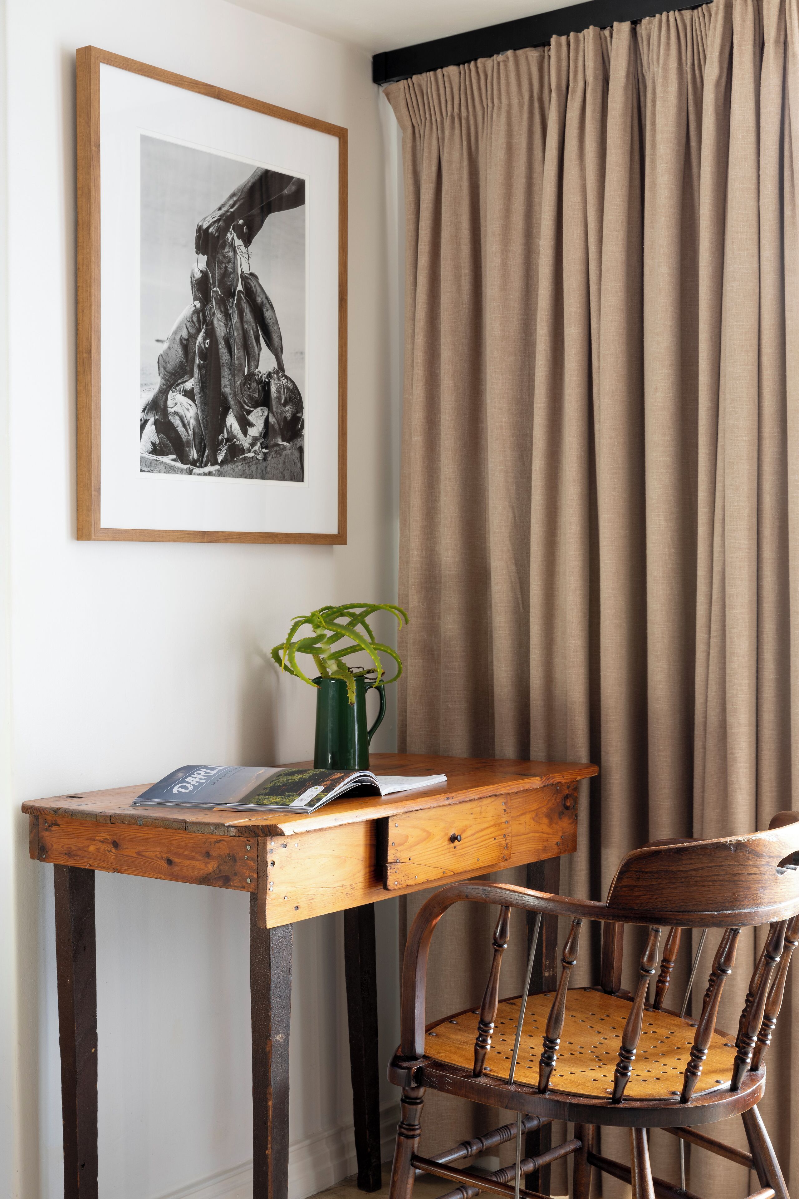 A wooden desk with a vase and a magazine, a wooden chair, a framed picture on the wall, and a curtain.