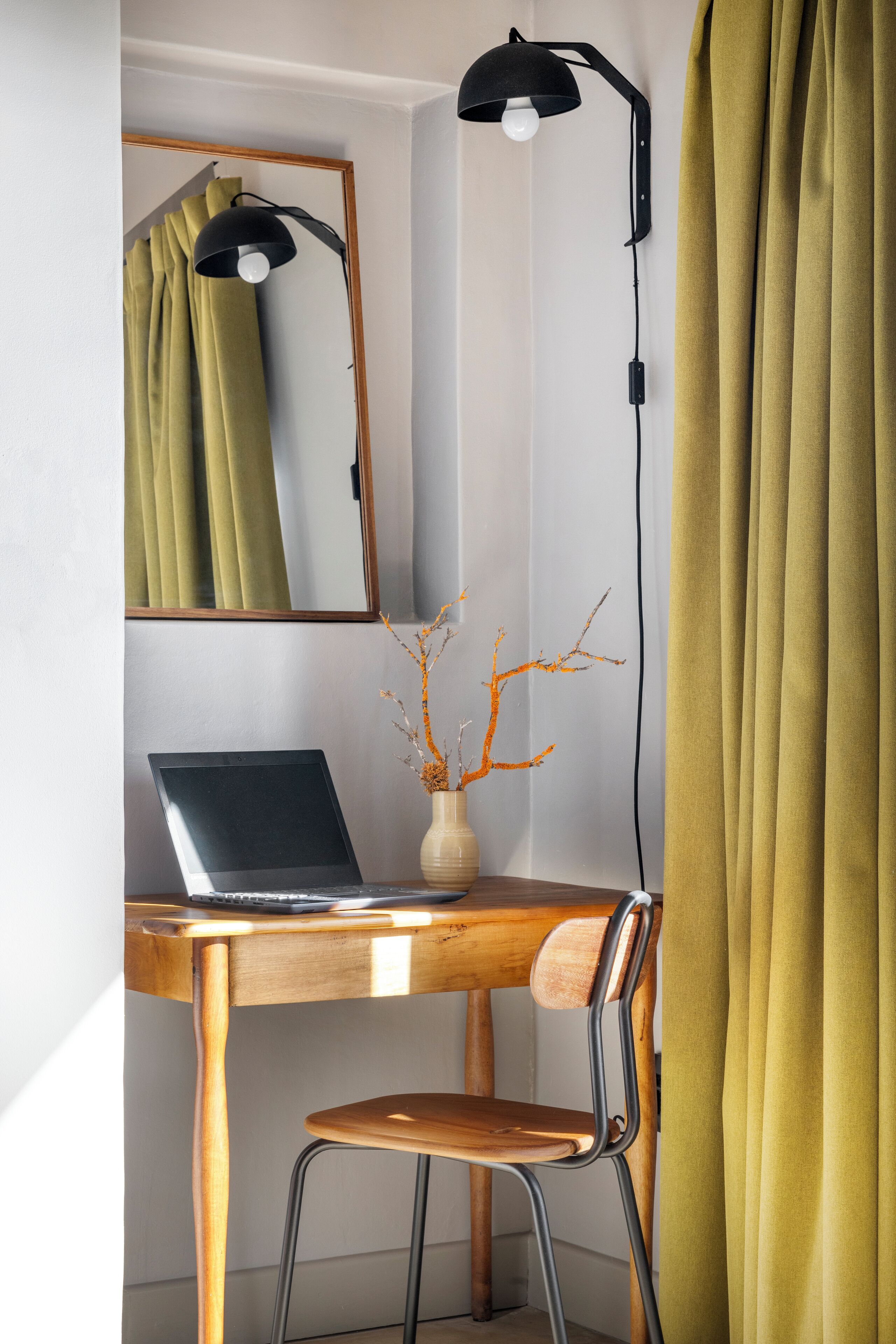 Un escritorio de madera con una laptop, un florero con ramas, una silla, una lámpara de escritorio y un espejo.