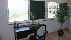 A room with a desk, chair, potted plant, and a view of the sea through large windows.