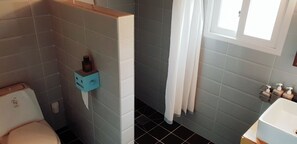 Une salle de bain avec des carreaux gris, des toilettes, un porte-savon bleu, une fenêtre avec des rideaux blancs et un lavabo avec des distributeurs de savon.
