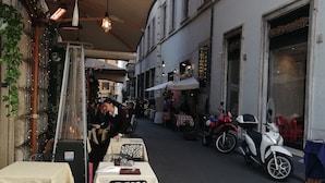 Un espace extérieur aménagé pour les repas, avec des tables et des chaises, une vue sur la rue où sont stationnés des scooters, et des bâtiments avec des enseignes.
