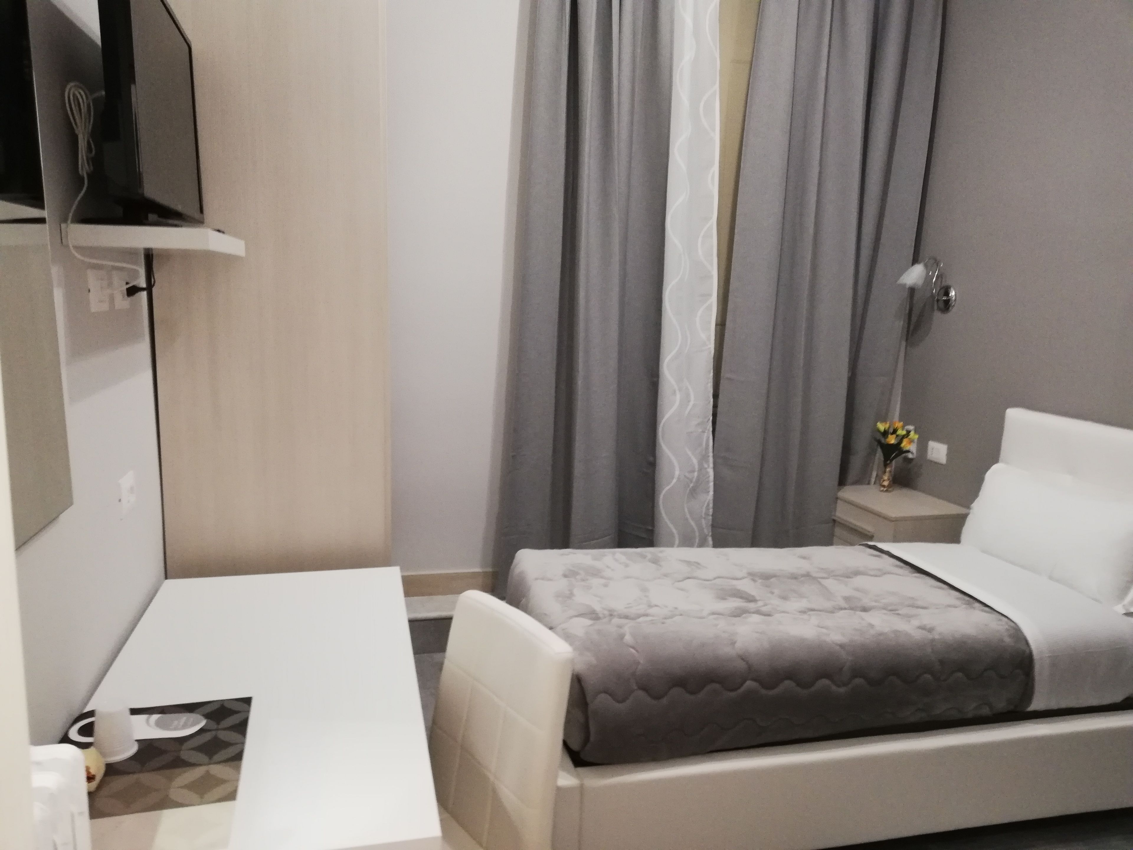 A hotel room with a bed, a desk, a chair, a TV, and a nightstand with a vase of flowers.