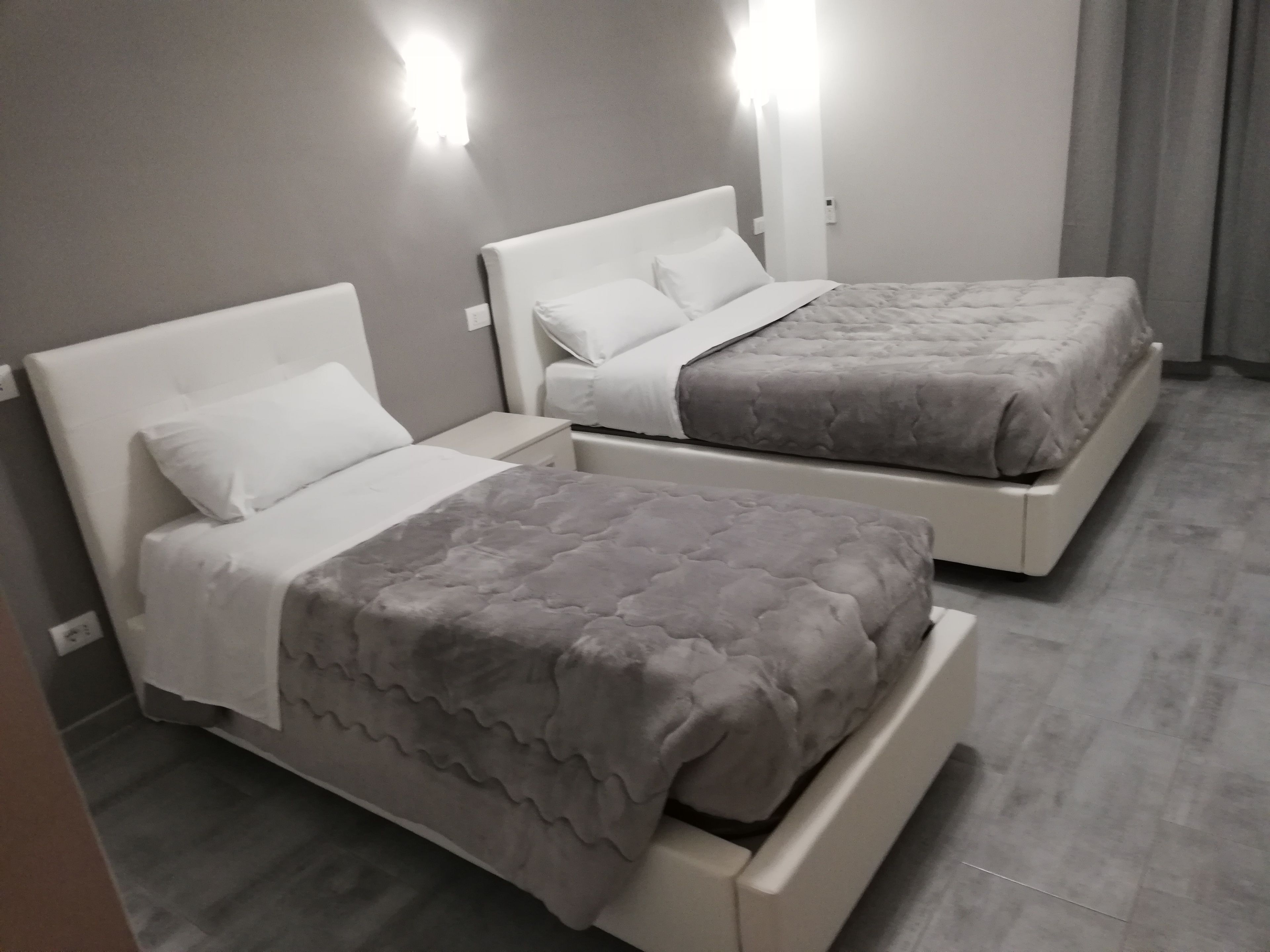 Une chambre d’hôtel avec deux lits, chacun doté d’une tête de lit blanche et d’une couvre-lit matelassée grise. Deux appliques murales sont fixées au-dessus de chaque lit.