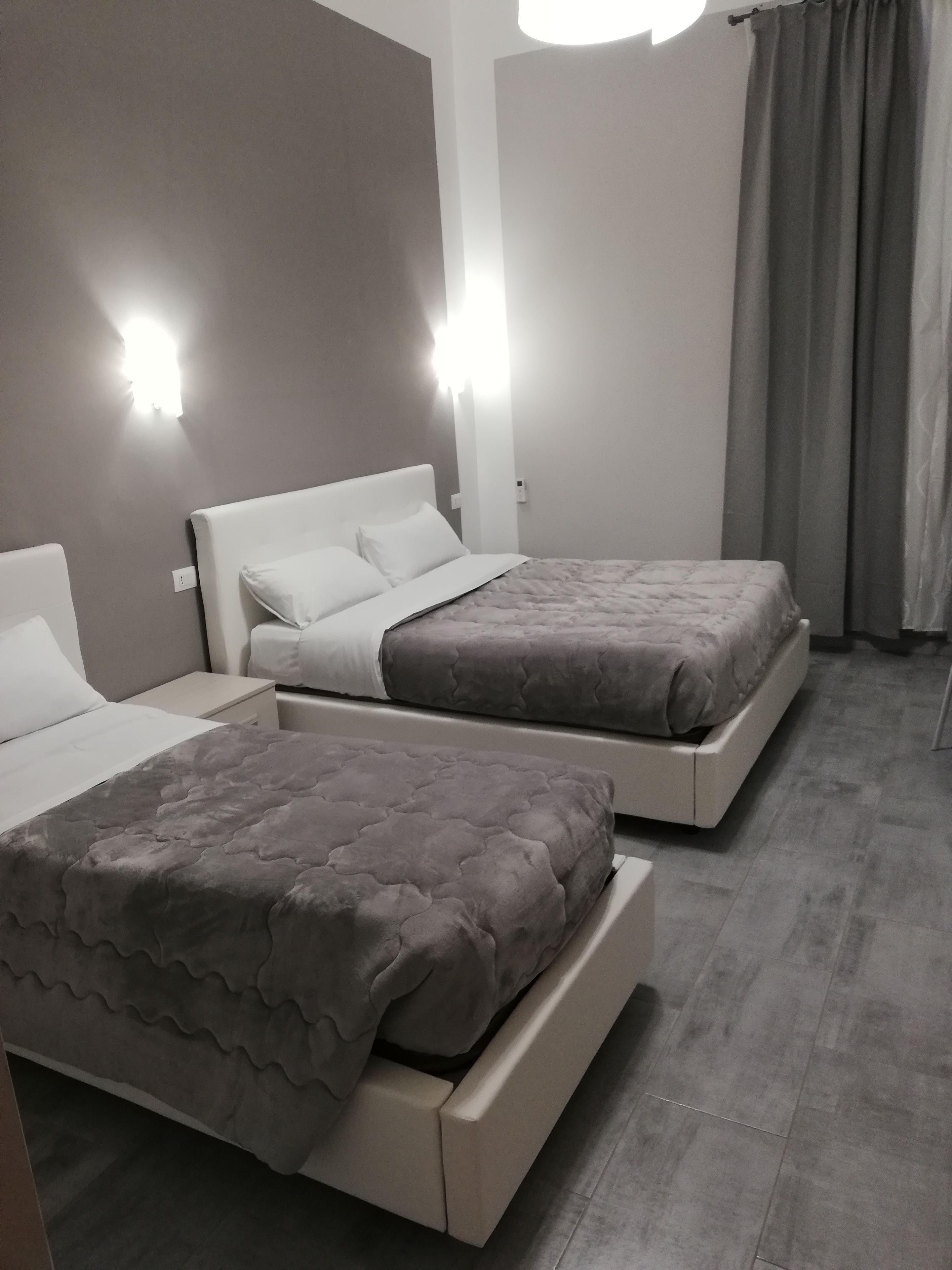 A hotel room with two beds, each with a grey quilted bedspread and white pillows. There are wall-mounted lights on either side of the beds, and a grey curtain on the window.