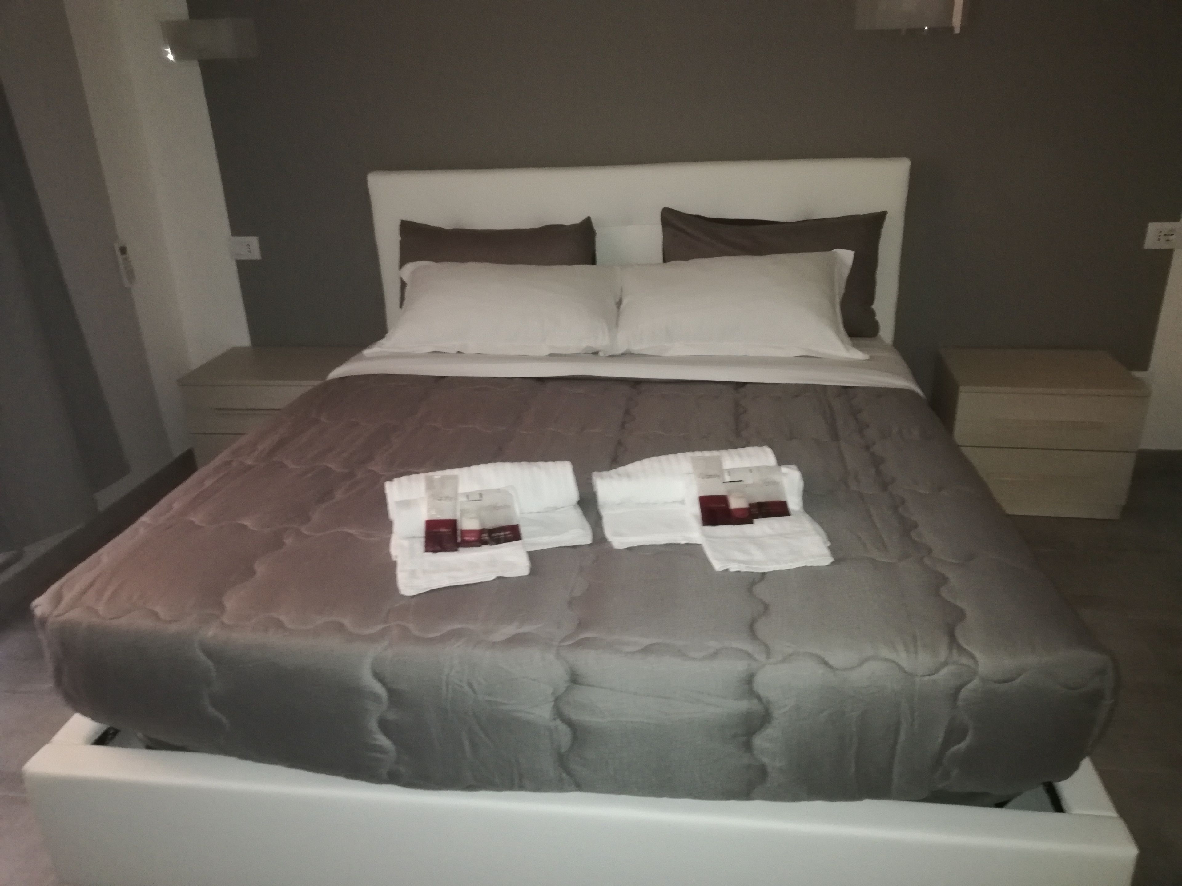 A neatly made bed with a grey duvet, two white pillows, and a folded towel with toiletries on it.