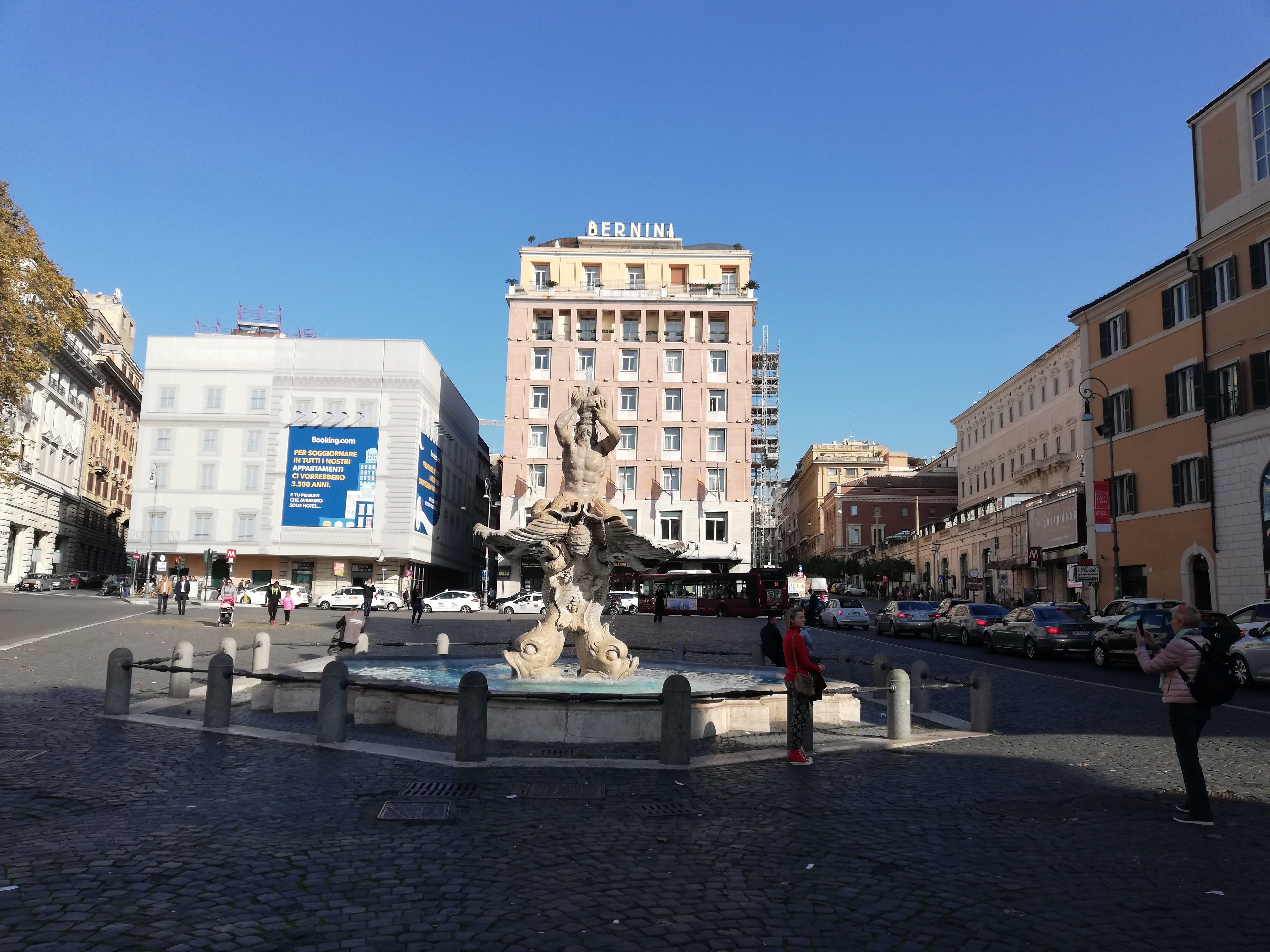 Une place publique avec une fontaine, entourée de bâtiments historiques et d’un hôtel reconnaissable à son grand panneau.